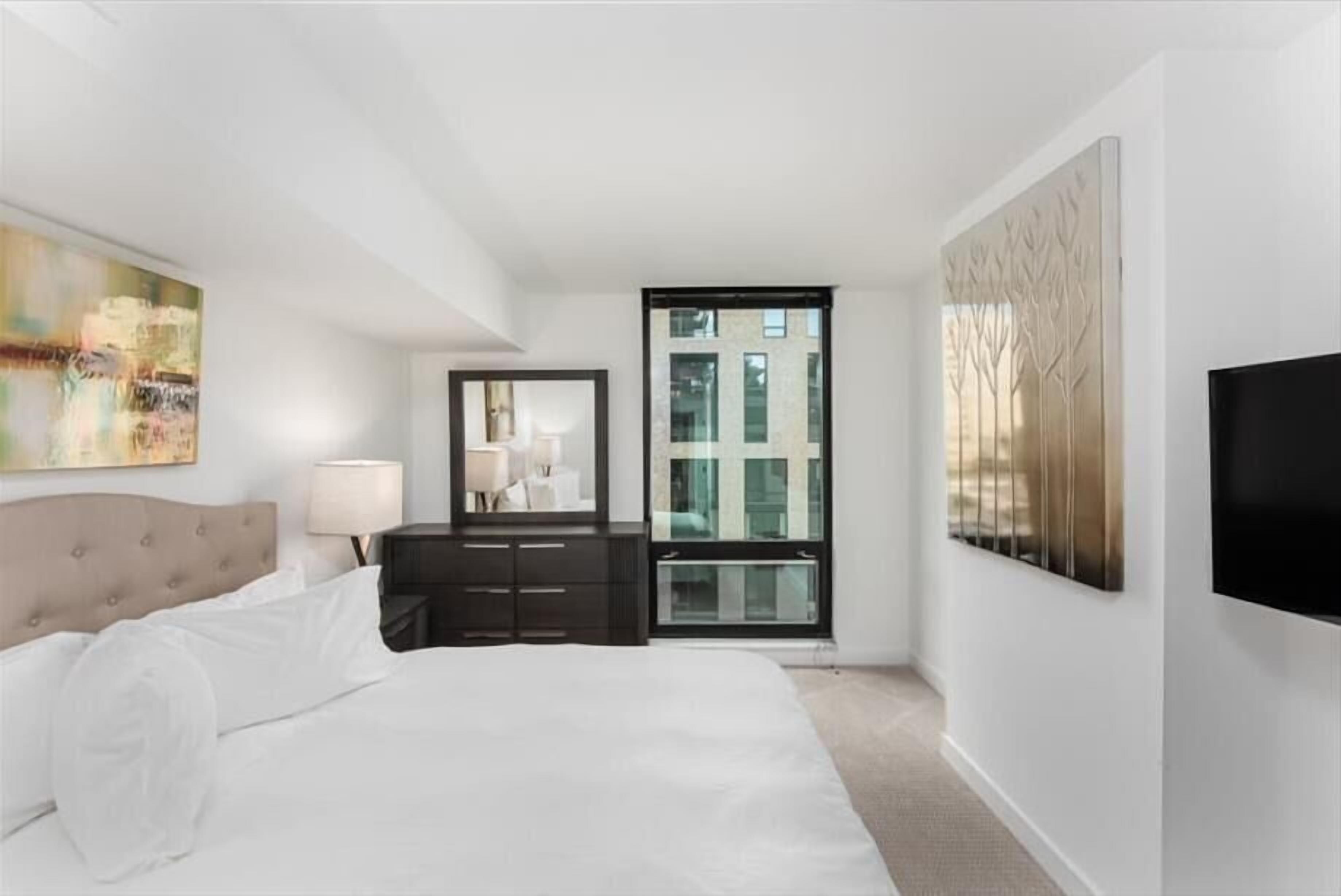 A modern hotel room with a large bed, a dresser, a mirror, a TV, and a window with a view of buildings.