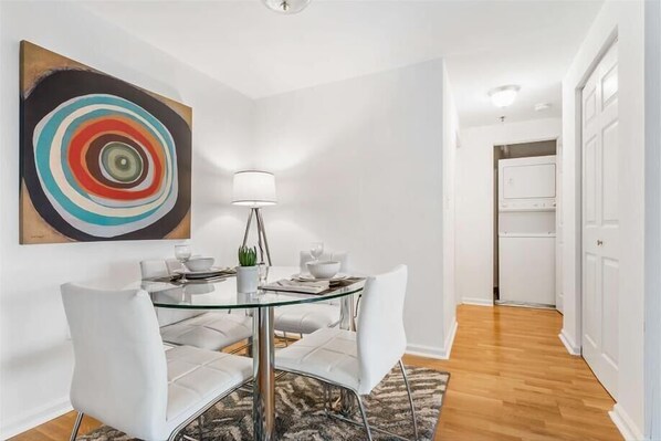 Un comedor con mesa de vidrio, sillas blancas, una lámpara moderna y un cuadro abstracto de colores en la pared.