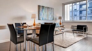 Un comedor moderno con una mesa redonda, sillas negras y una superficie de vidrio. Hay un cuadro en la pared y una ventana con vistas a edificios.