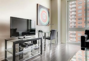 A modern living room with a flat-screen TV on a glass console, a glass table, a black chair, and a large window with a view of a red building. - Spacious & Clean 1BR | Work Desk & WiFi | by GLS (Jersey City)
