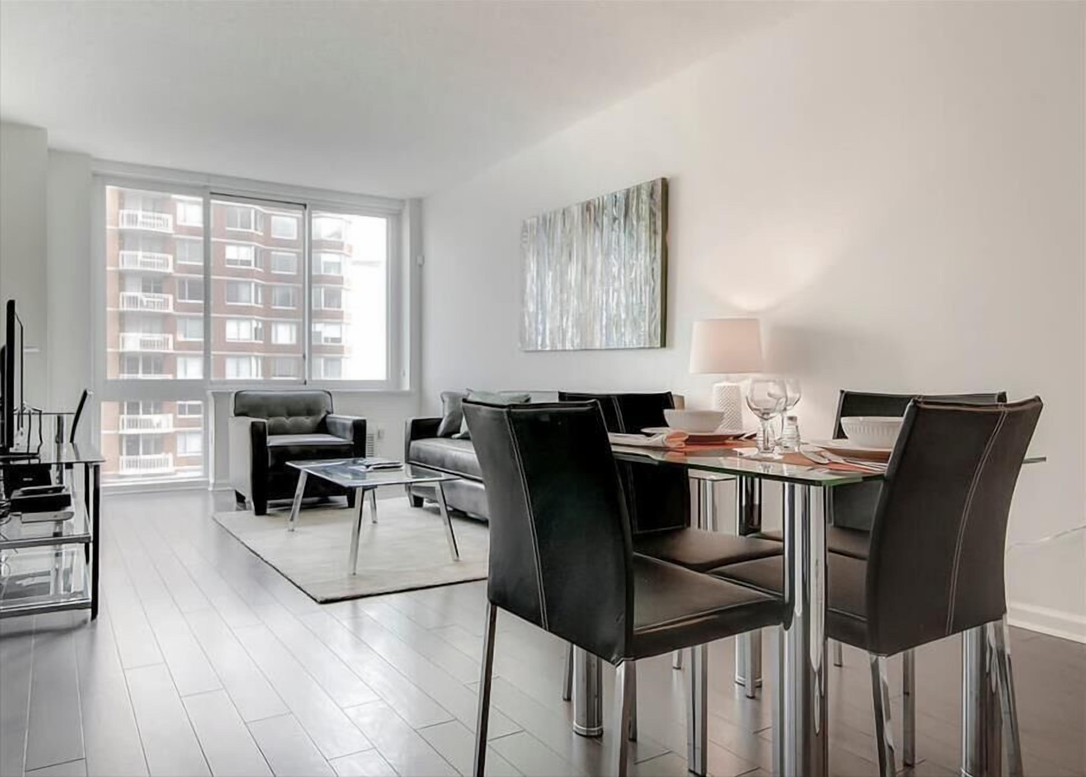 A modern living room with a dining area, featuring a sofa, chairs, a glass table, and a view of a building through large windows.