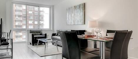 A modern living room with a dining area, featuring a sofa, chairs, a glass table, and a view of a building through large windows.