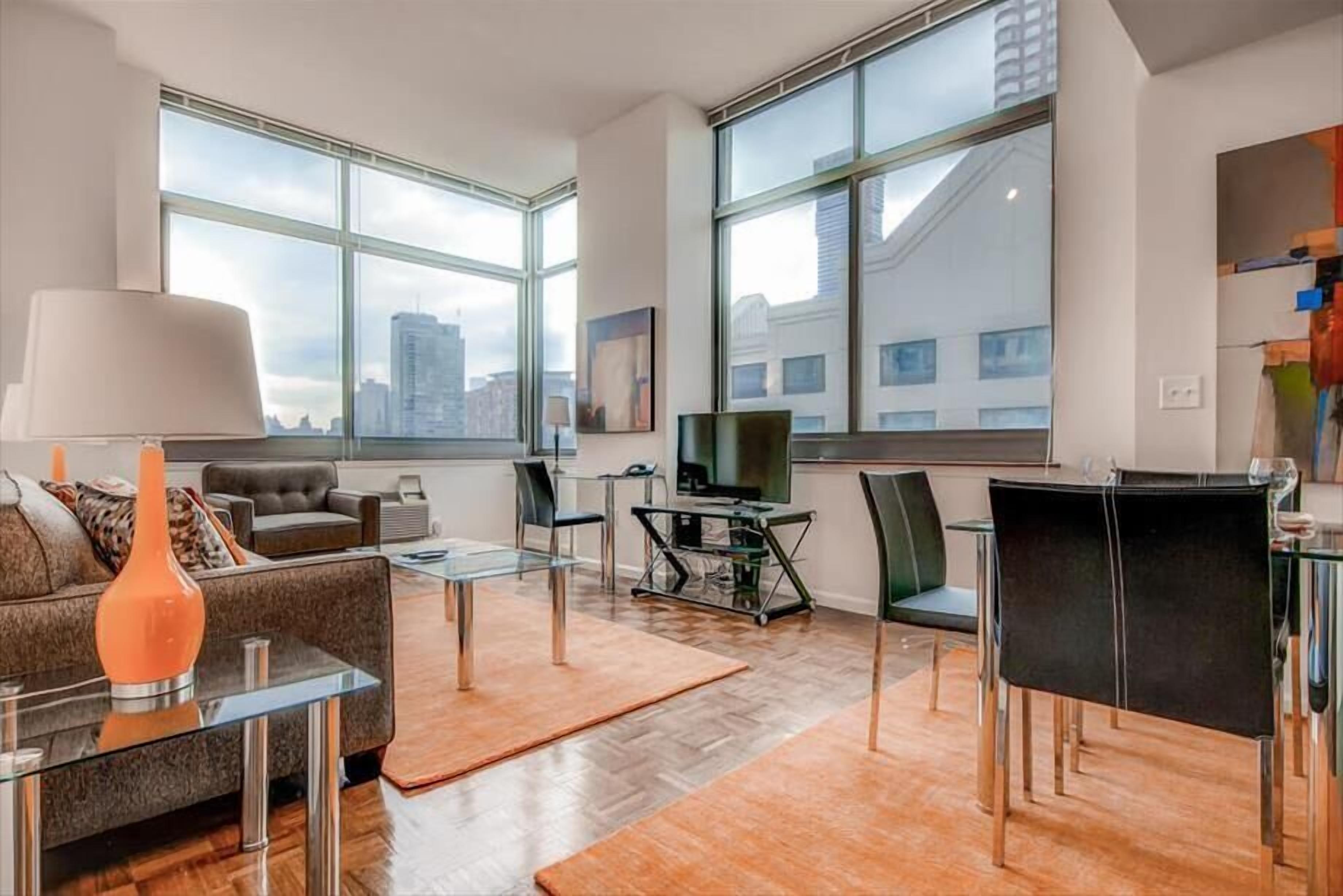 Una sala de estar moderna con sofá, mesa de centro y televisor, ventanas amplias con vista a la ciudad y un comedor con sillas y mesa.