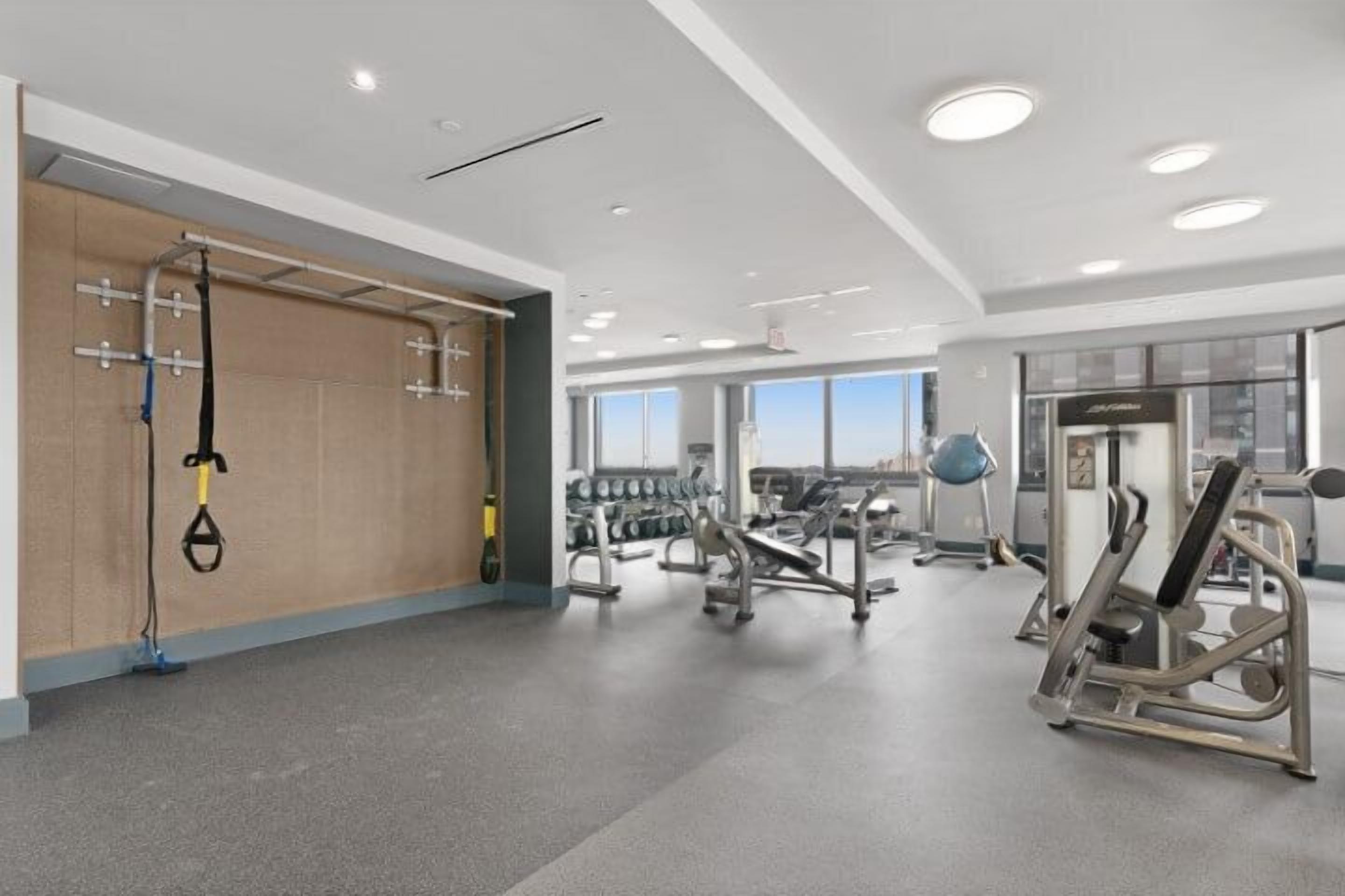A modern gym with various exercise equipment, large windows, and a high ceiling.