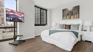 Ein modernes Schlafzimmer mit einem großen Bett, einem Fernseher, einem Fenster mit Blick auf die Stadt und einem Bild an der Wand.