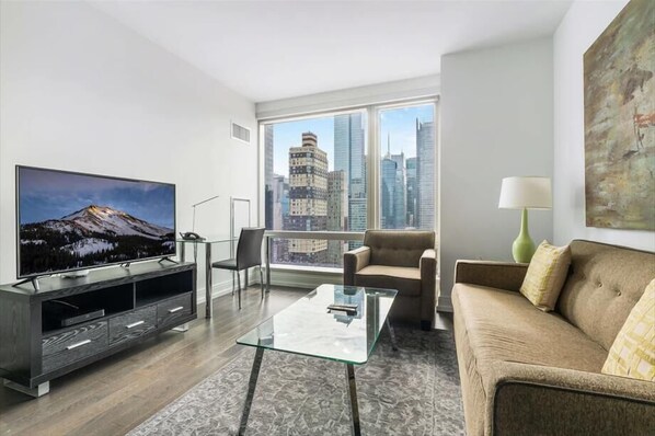 A modern living room with a large TV, a glass coffee table, a sofa, and a view of city buildings.