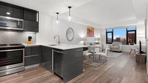 Una cocina moderna con microondas integrado, horno y una isla con lavamanos. Hay un comedor con mesa y sillas, y un ventanal con vistas a la ciudad.