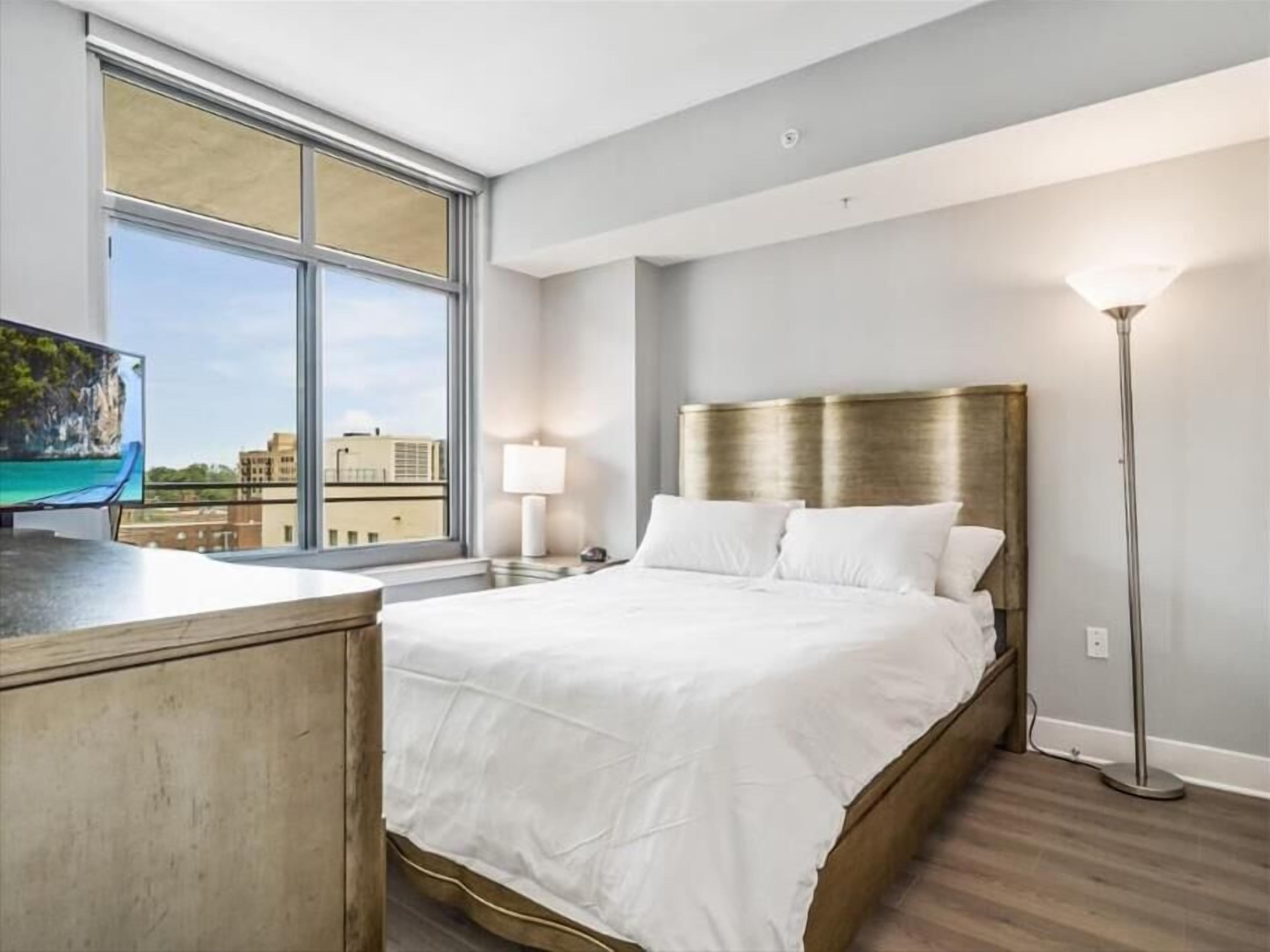 Une chambre d’hôtel moderne avec une grande fenêtre, un lit doté d’une tête de lit dorée, une table de chevet et un lampadaire.