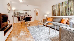Ein modernes Wohnzimmer mit Sofa, Couchtisch und Fernseher. Im Hintergrund befindet sich eine Küche mit Essbereich.