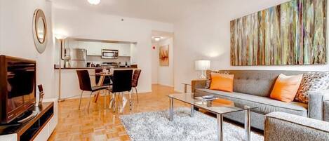 Una sala de estar moderna con sofá, mesa de centro y televisor. Se ve una cocina al fondo con una zona de comedor.