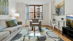 Ein modernes Wohnzimmer mit Glastisch, Sofa, Sessel, Fernseher und Kunstwerken an den Wänden.