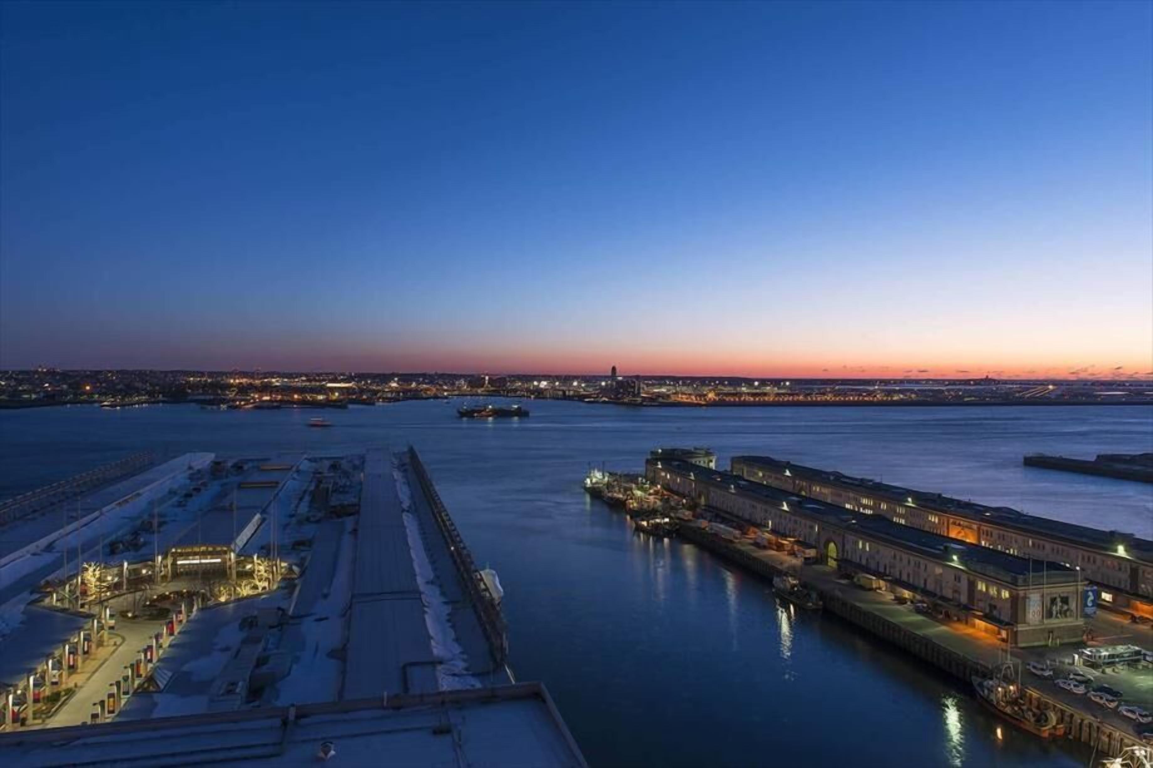 A waterfront cityscape at dusk with illuminated buildings and a calm river.