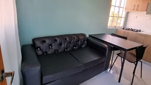 Un espace de vie compact comprenant un canapé en cuir noir, une petite table à manger et un coin cuisine avec des meubles en bois et une cuisinière à gaz.