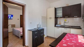 A compact hotel room with a kitchenette, a bed with a checkered blanket, a small desk with a kettle, and a TV in the corner.