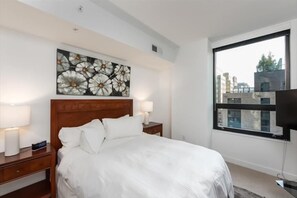 Ett hotellrum med en stor säng, två sängbord med lampor, ett konstverk som hänger på väggen och ett stort fönster med utsikt över byggnader.