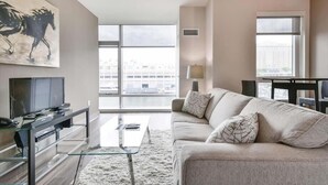Un soggiorno moderno con una grande finestra, una TV a schermo piatto, un tavolino da caffè in vetro e un divano color beige.