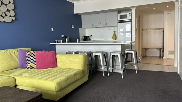 Un salon moderne avec un canapé jaune, une table basse et un coin cuisine comprenant un comptoir blanc, des tabourets en acier inoxydable et un four à micro-ondes.