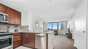 A modern kitchen with a microwave, stove, and dishwasher, a large island with a sink, and a view of the city through large windows. - Spacious 1BR | Gym | Rooftop Pool | Downtown Jersey City | GLS (Jersey City)