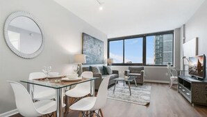 A modern living room with a glass dining table, white chairs, a sofa, and a large window. - Spacious 1BR | Gym | Rooftop Pool | Downtown Jersey City | GLS (Jersey City)