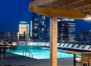 A rooftop pool with lounge chairs, a wooden pergola, and a city skyline view at night. - Modern 1BR | Full Kitchen | W/D |  NYC Views | by GLS (Jersey City)
