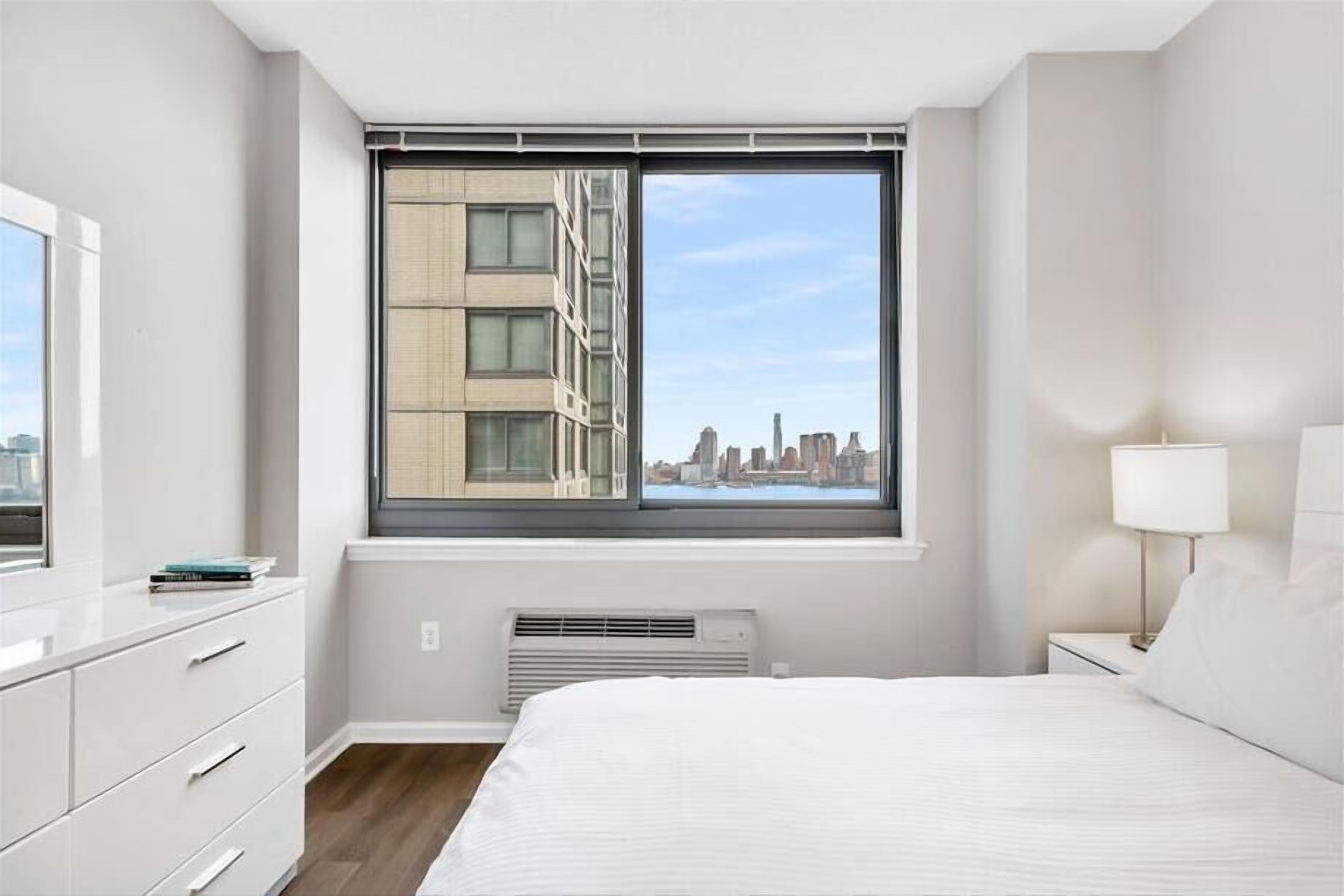 A modern bedroom with a large window, a bed, a bedside table with a lamp, and a white dresser.