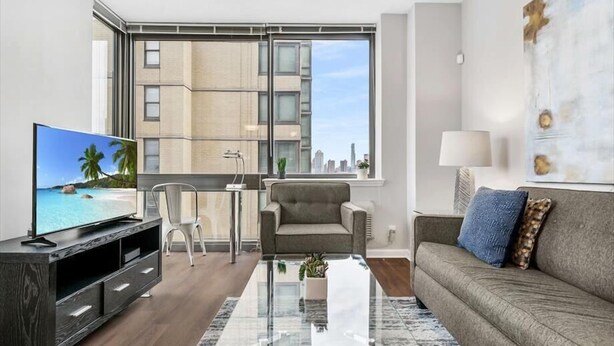 A modern living room with a large TV, a sofa, a coffee table, and a view of a cityscape through large windows.
