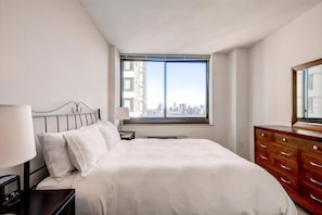 A hotel room with a large bed, a nightstand with a lamp, a dresser, and a window with a city view.