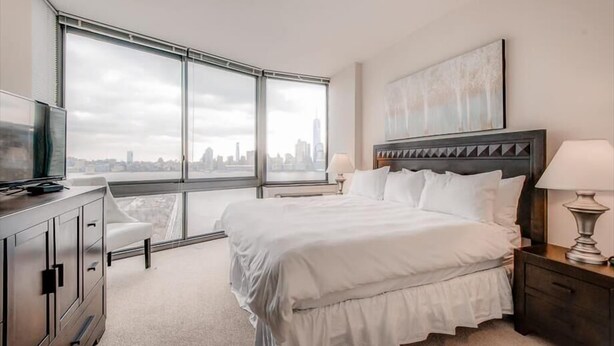 A hotel room with a large window, a bed with white bedding, a bedside table with a lamp, and a television on a cabinet. - Spacious 2BR | Fully Equipped Gym | Near PATH by GLS (Jersey City)
