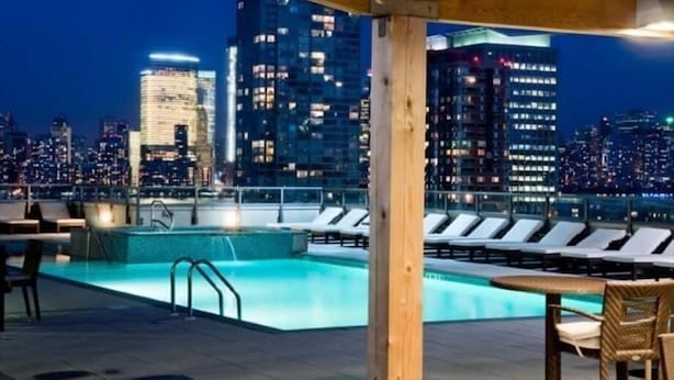 A rooftop pool with lounge chairs, a wooden pergola, and a city skyline view at night. - Spacious 1BR | Full Kitchen | Near PATH by GLS (Jersey City)