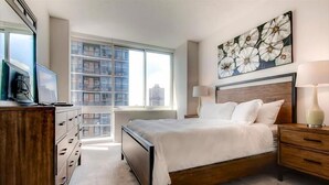 A modern hotel room with a large bed, a desk with a computer, a dresser, and a window with a city view. - Modern 1 Bedroom Apartments with Manhattan views (Jersey City)