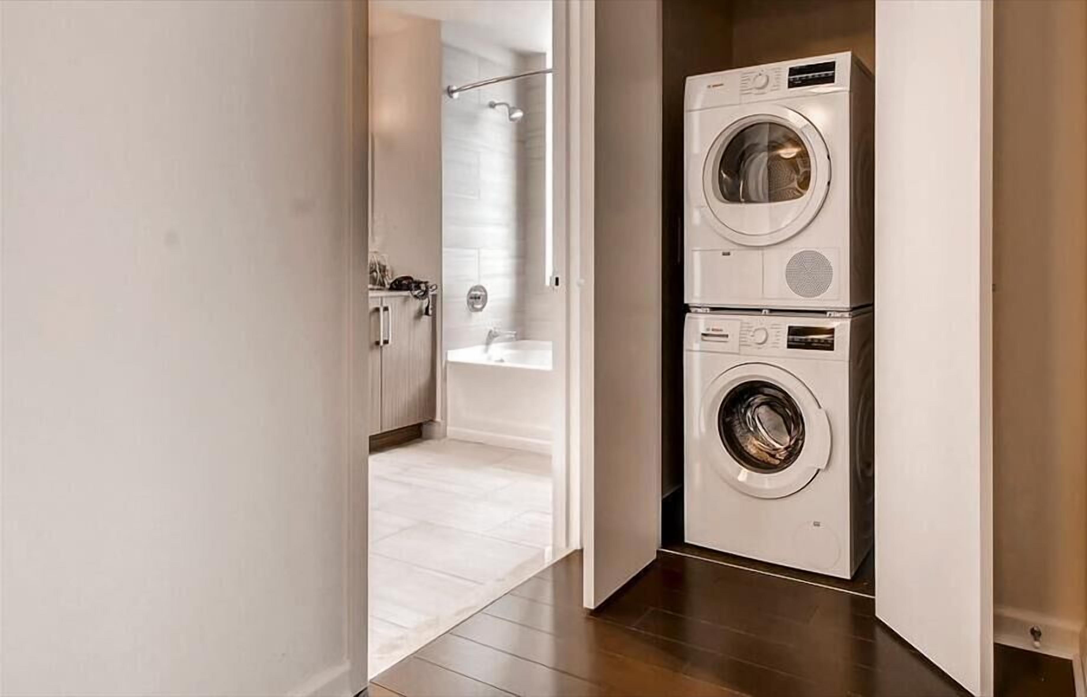 Un cuarto de lavandería con dos lavadoras de carga frontal apiladas y una zona de ducha visible a través de una puerta abierta.