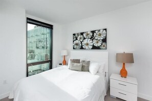 A modern bedroom with a large bed, two bedside tables with lamps, a window with blinds, and a floral artwork on the wall.