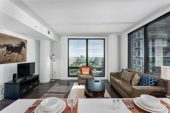 A modern living room with a large window, a flat-screen TV, a sofa, and a dining table set for two.