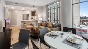 A modern living room with a dining area, a kitchen in the background, and large windows offering a view of the city.