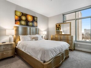 Ein Hotelzimmer mit einem großen Bett, zwei Nachttischen, einer Kommode, einem Spiegel und einem großen Fenster mit Blick auf die Stadt.