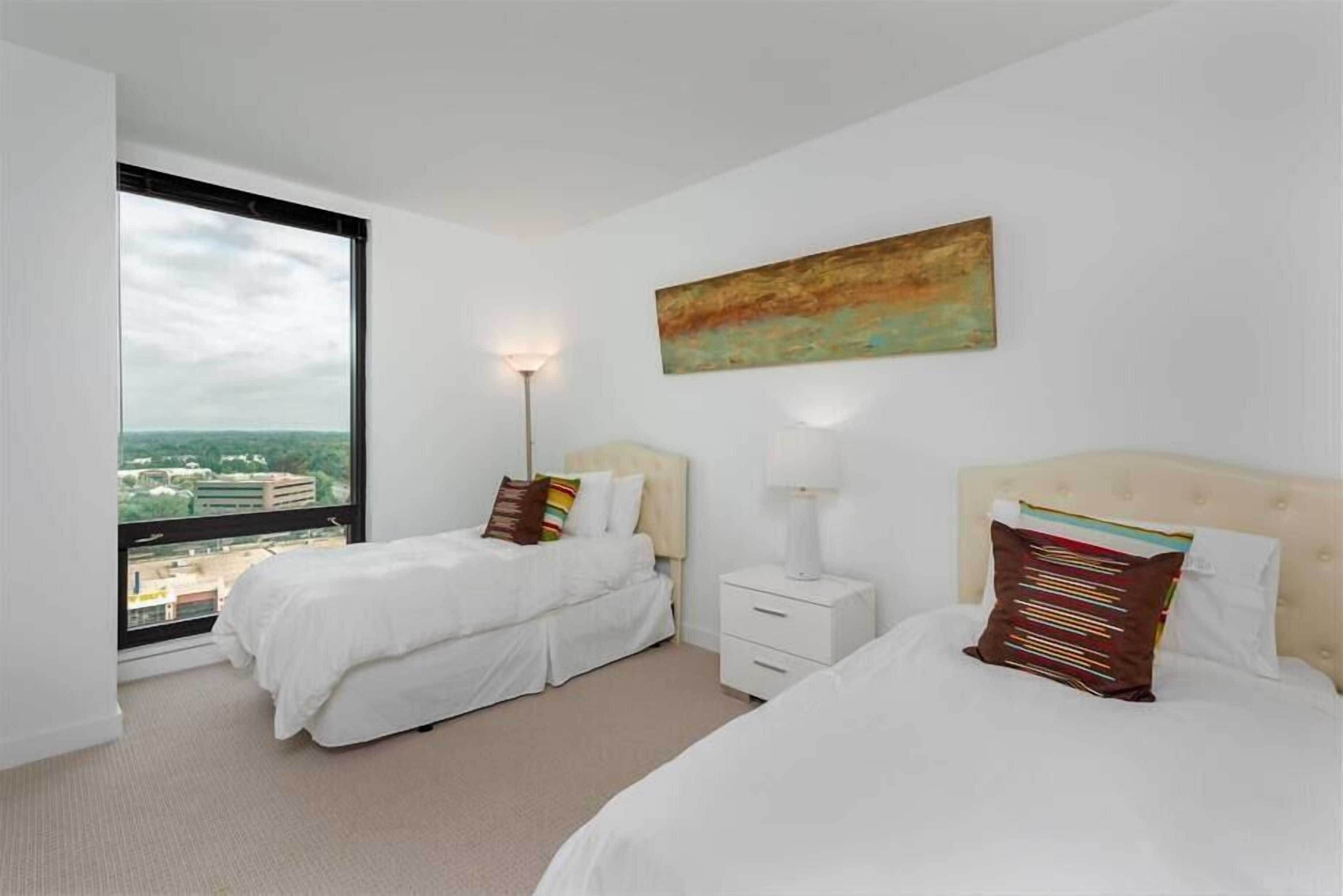 Een hotelkamer met twee bedden, een groot raam, een schilderij aan de muur en een nachtkastje met een lamp.