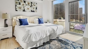 A modern bedroom with a large bed, white bedding, a blue and white patterned rug, a nightstand with a lamp, and a large window with a city view. - Arlington Virginia 2BR | Crystal City | Easy DC Access | by GLS (Arlington)