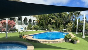 A swimming pool with a surrounding green lawn, a covered area with a roof, and a building with arched windows in the background. - Magnetic Island Resort Studio - Relax, sun, swim, fish, beach and lovely gardens (Nelly Bay)