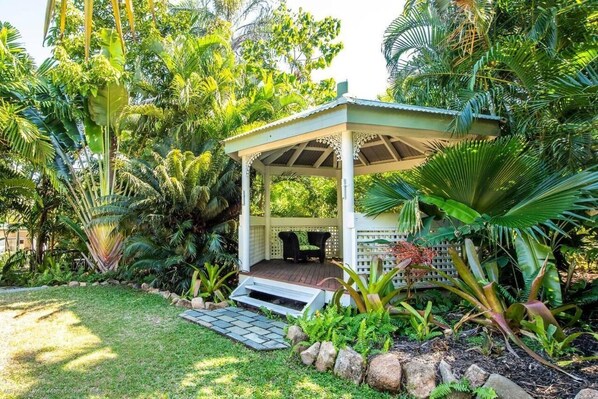 A gazebo with a wooden deck and a white lattice roof, surrounded by lush tropical vegetation.