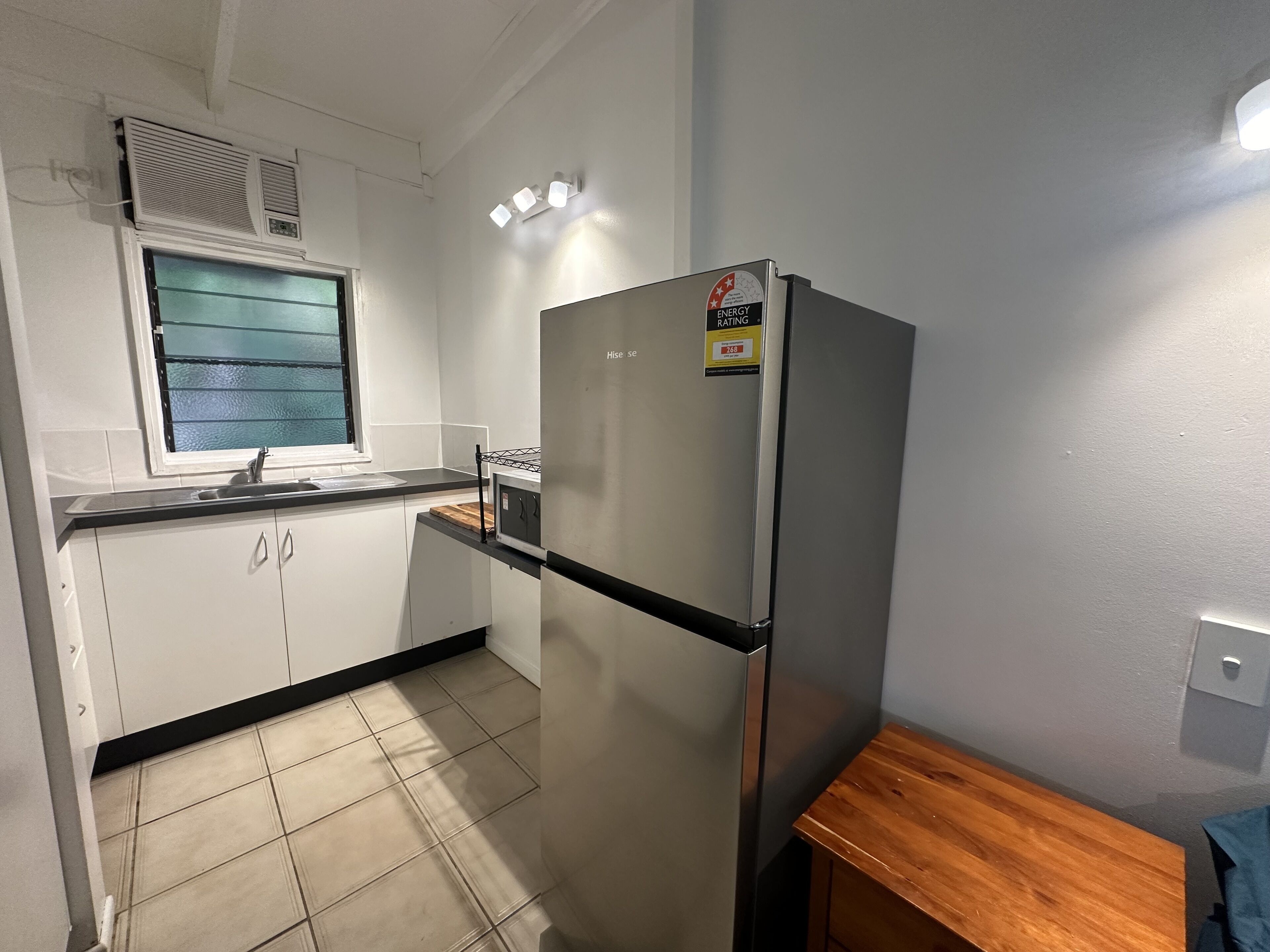 A compact kitchen with white cabinets, a stainless steel refrigerator, a window with blinds, and a wooden countertop.