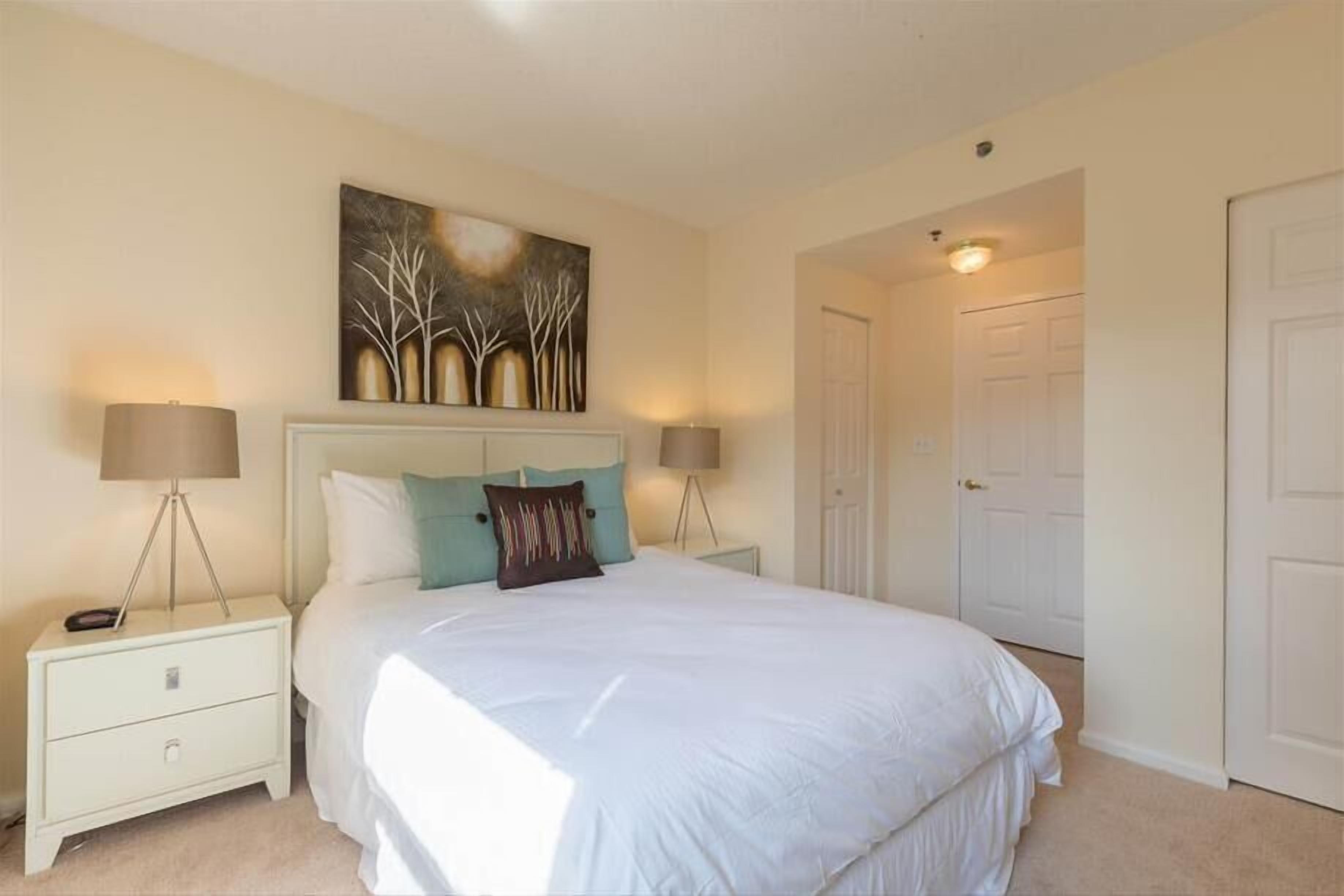 A bedroom with a large bed, two bedside tables with lamps, a painting on the wall, and a door to the right.