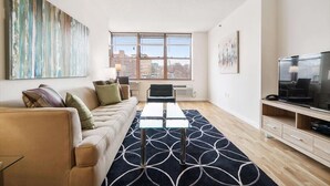 A modern living room with a sofa, a glass coffee table, a TV, and a large window with a city view. - Private & Clean 1BR | Work Remote | Hoboken Riverfront | Easy access to NYC (Hoboken)