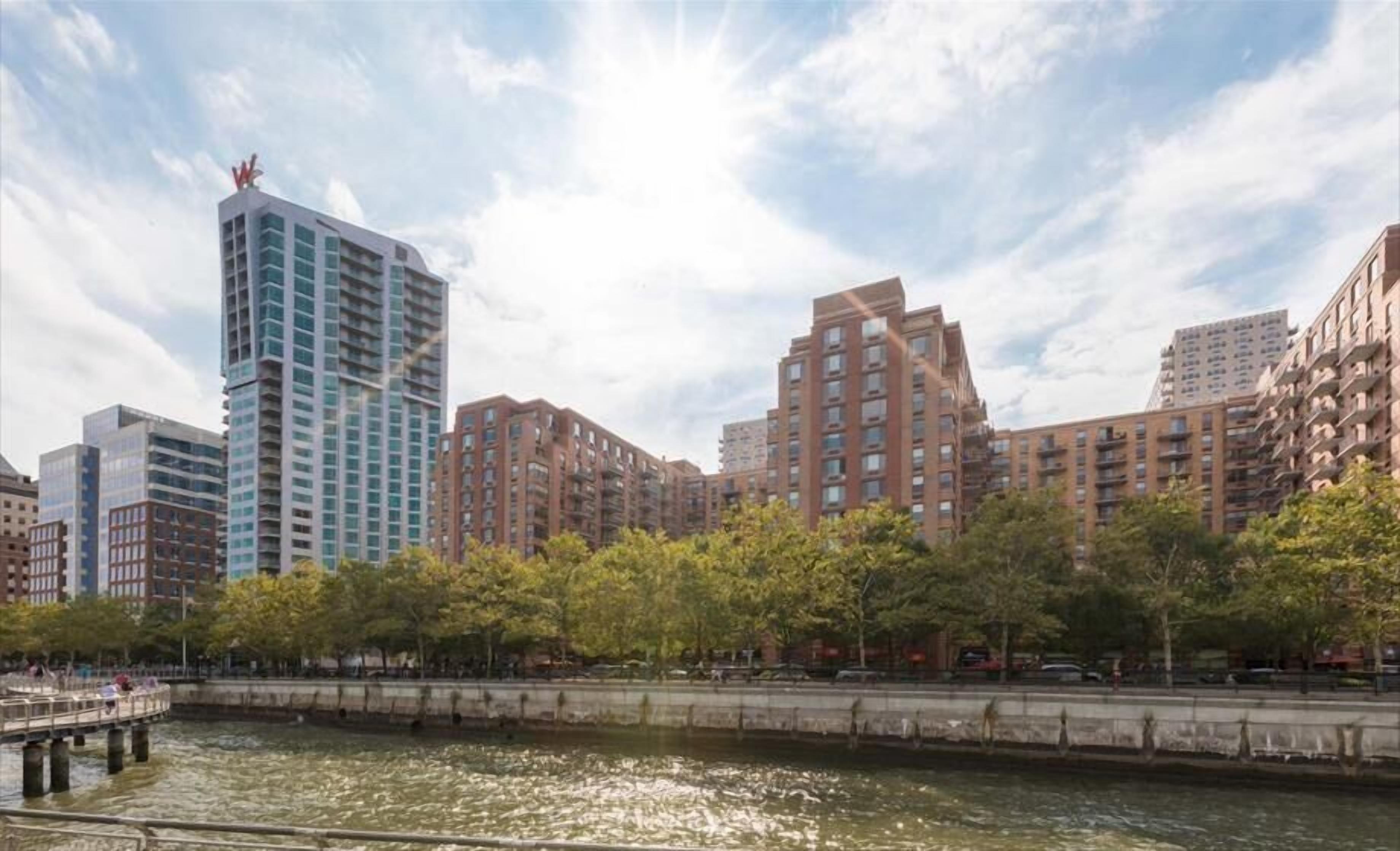 A riverside urban area with modern high-rise buildings, a bridge, and a park with trees.