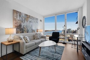 A modern living room with a large window, a sofa, a coffee table, and a TV. - Spacious 1BR | Full Kitchen | Near Javits Center | by GLS (New York)