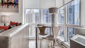 Una sala de estar moderna con una mesa de vidrio, un sofá y un sillón. Se ve una ventana grande con vistas a edificios altos.