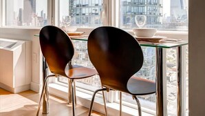 Un espace repas moderne, composé de deux chaises et d’une table en verre, offrant une vue sur un paysage urbain avec de hauts immeubles.