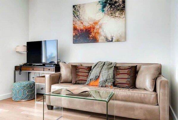 Un salon moderne avec un canapé en cuir beige, une table basse en verre, un téléviseur à écran plat posé sur un meuble en bois et un tableau abstrait aux couleurs vives accroché au mur.