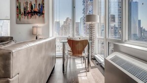 Un salon moderne doté d’une grande fenêtre, offrant une vue sur la ville, avec un canapé, une table à manger et des chaises, ainsi qu’un lampadaire.