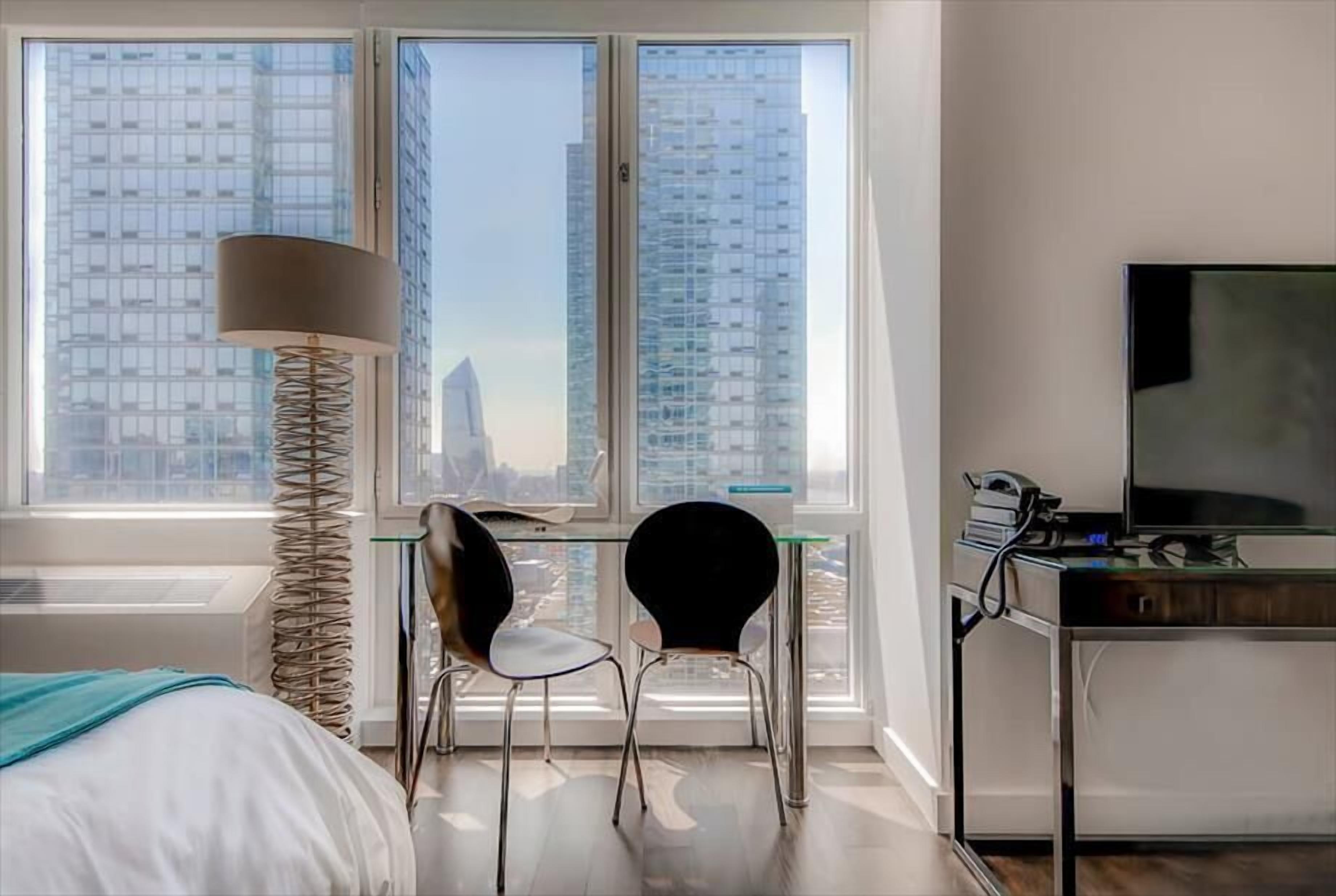 Ein modernes Hotelzimmer mit einem Bett, einem Schreibtisch mit zwei Stühlen, einer Lampe, einem Fernseher und einem großen Fenster mit Blick auf die Stadt.