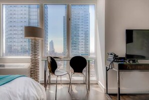 A modern hotel room with a bed, a desk with two chairs, a lamp, a TV, and a large window with a city view.